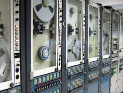 The machines pictured read the inch-wide magnetic data tape from their 14-inch reels. Multiple machines are used because each reel only records about 15 minutes worth of data. As one reel fills, the next machine automatically starts recording a slight overlap for data continuity.